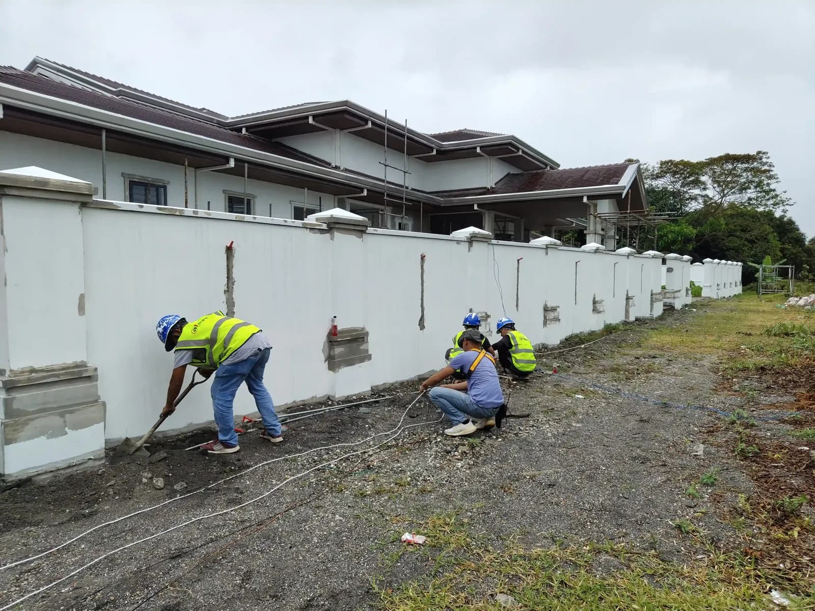A crew in hi-vis vests trenching and laying cable along a perimeter wall.